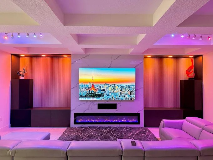Media room with a wide television wall, linear fireplace and colour-changing cove lighting across a coffered ceiling.